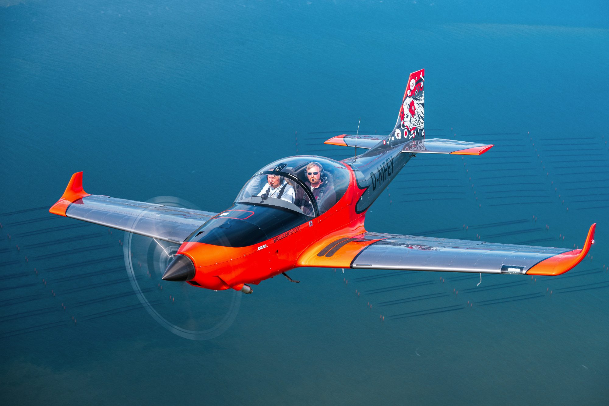 Air-to-Air Luftbild einer Breezer Sport über der Nordsee Ein rotes und schwarzes Sportflugzeug fliegt über einem stillen Gewässer.