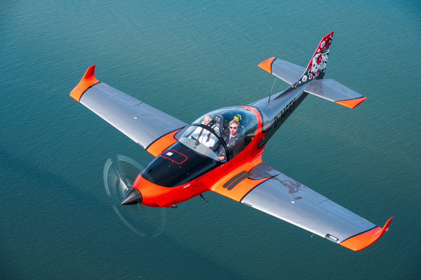 Ein oranges und graues Flugzeug fliegt über blauen Wasserfläche.