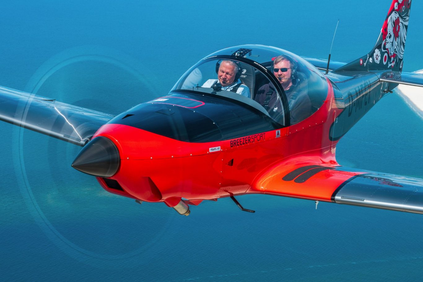 Air-to-Air Luftbild einer Breezer Sport über der Nordsee Zwei Piloten sitzen in einem roten Sportflugzeug, das über blaues Wasser fliegt.